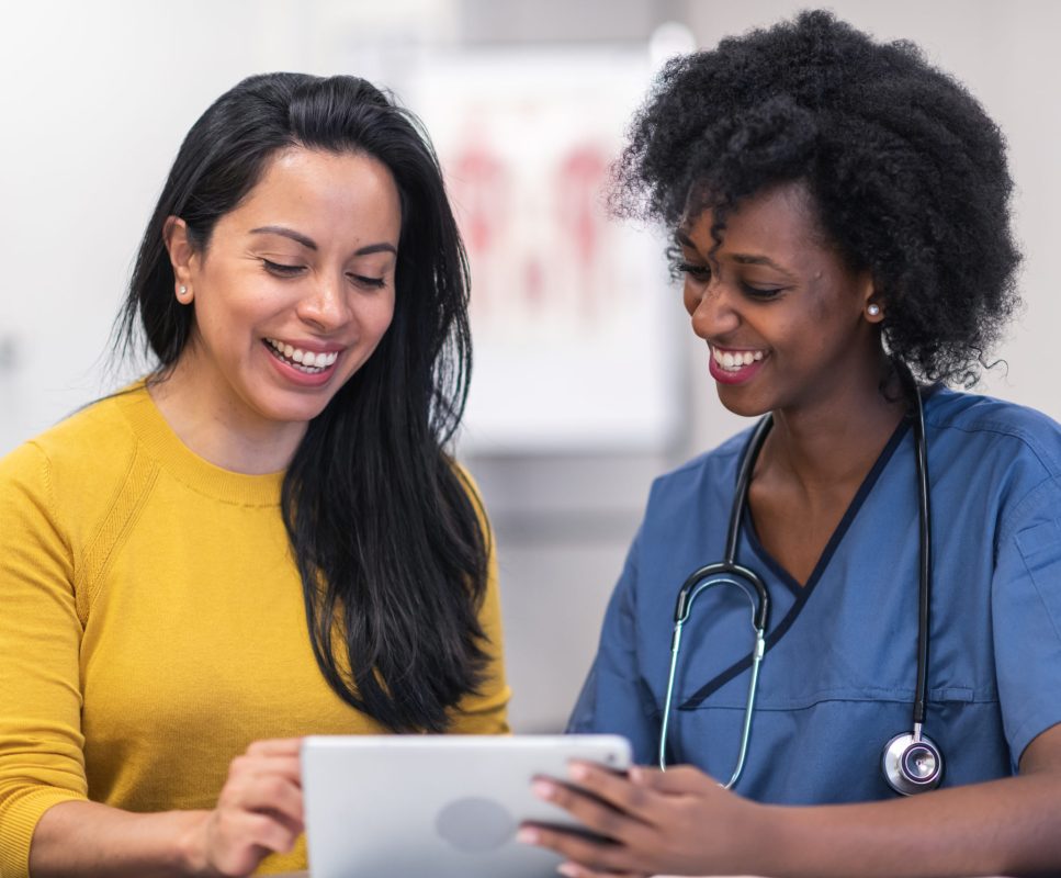 A woman of Hispanic descent is at a routine medical appointment. Her healthcare provider is a young black woman. The two women are seated at a table together in a medical examination room. The doctor is holding a digital tablet and is showing the patient test results. Both individuals are smiling.